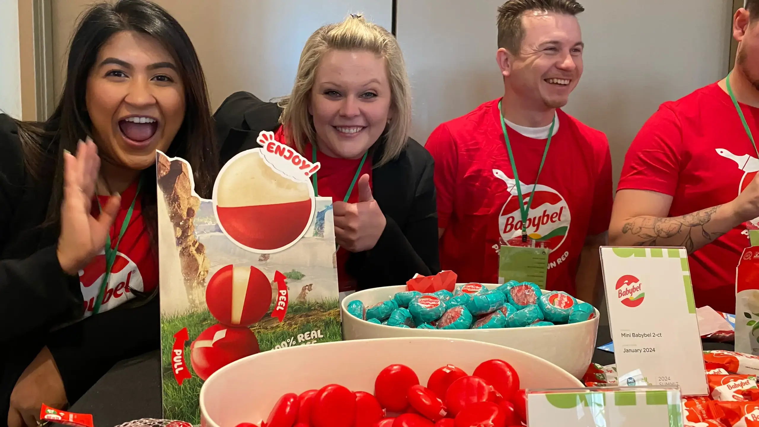 Three smiling brand ambassadors wearing red Babybel shirts stand behind a table of Babybel cheese samples and promotional signage at a conference booth