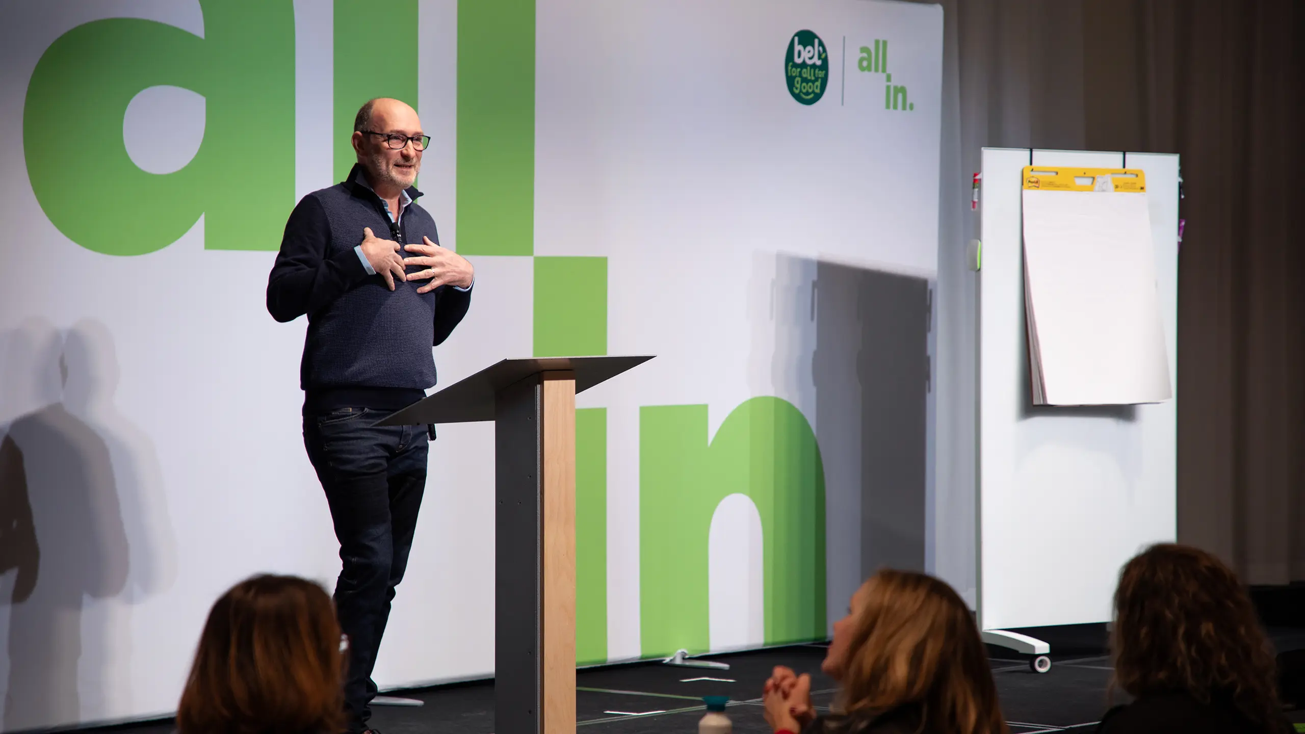 Speaker presenting at Bel Brands’ ‘All In’ leadership summit, standing at a podium in front of large green ‘all in’ branding