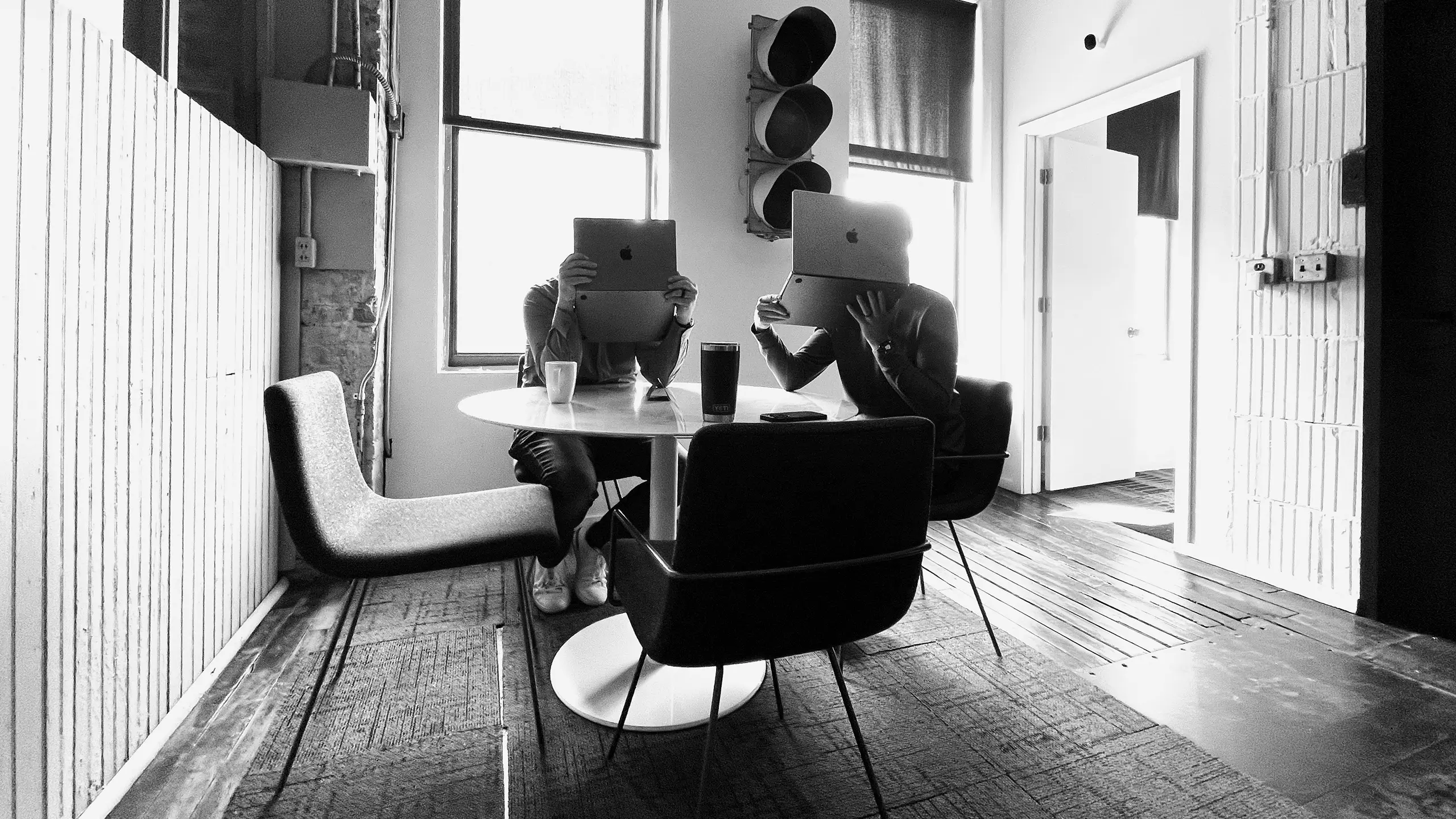 Multiple team members sitting at a round table in a bright office, holding laptops during a casual, collaborative work session.