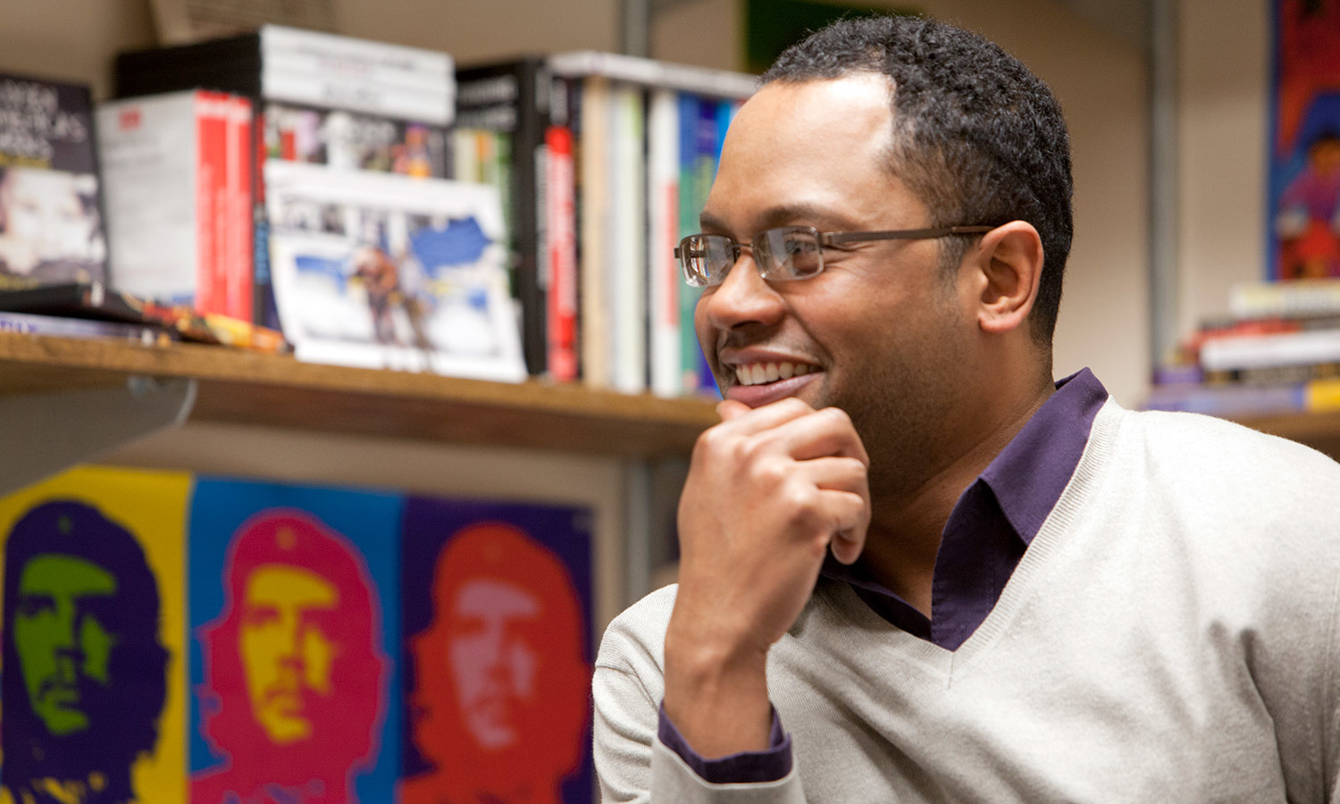 A man smiles thoughtfully in a cozy office. Books and vibrant pop art featuring are visible on the shelves behind him.