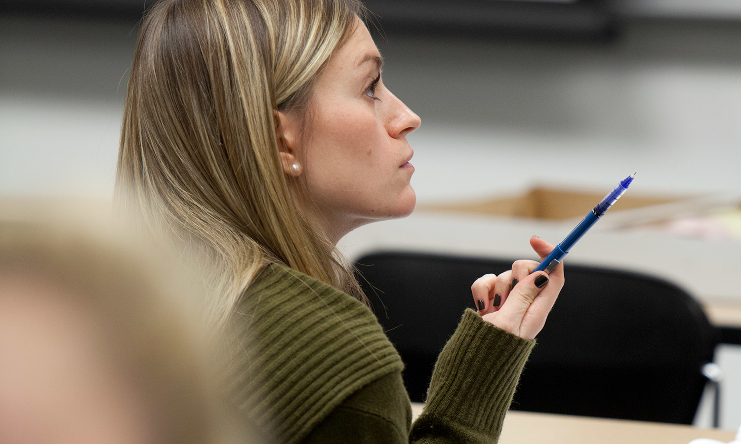 A woman sitting in a classroom, holding a blue pen, wearing a green sweater, conveying focus and thoughtfulness.
