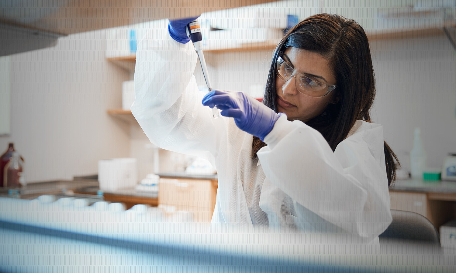 A scientist in a lab coat and safety goggles carefully uses a pipette in a laboratory. She appears focused and precise. Laboratory equipment is visible in the background.