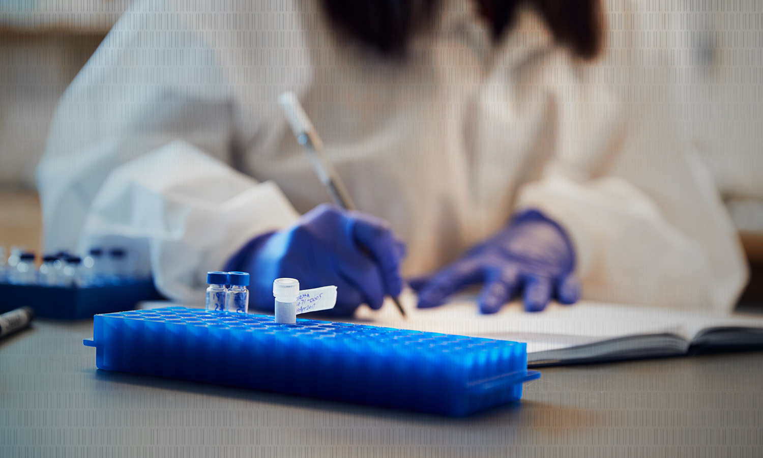 A scientist in a lab coat and blue gloves writes in a notebook. In the foreground, a blue tray holds vials, suggesting a focused research setting.