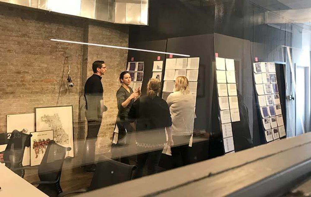 A team of four people engage in a discussion in Multiple's office with exposed brick walls and storyboard sheets pinned to walls.